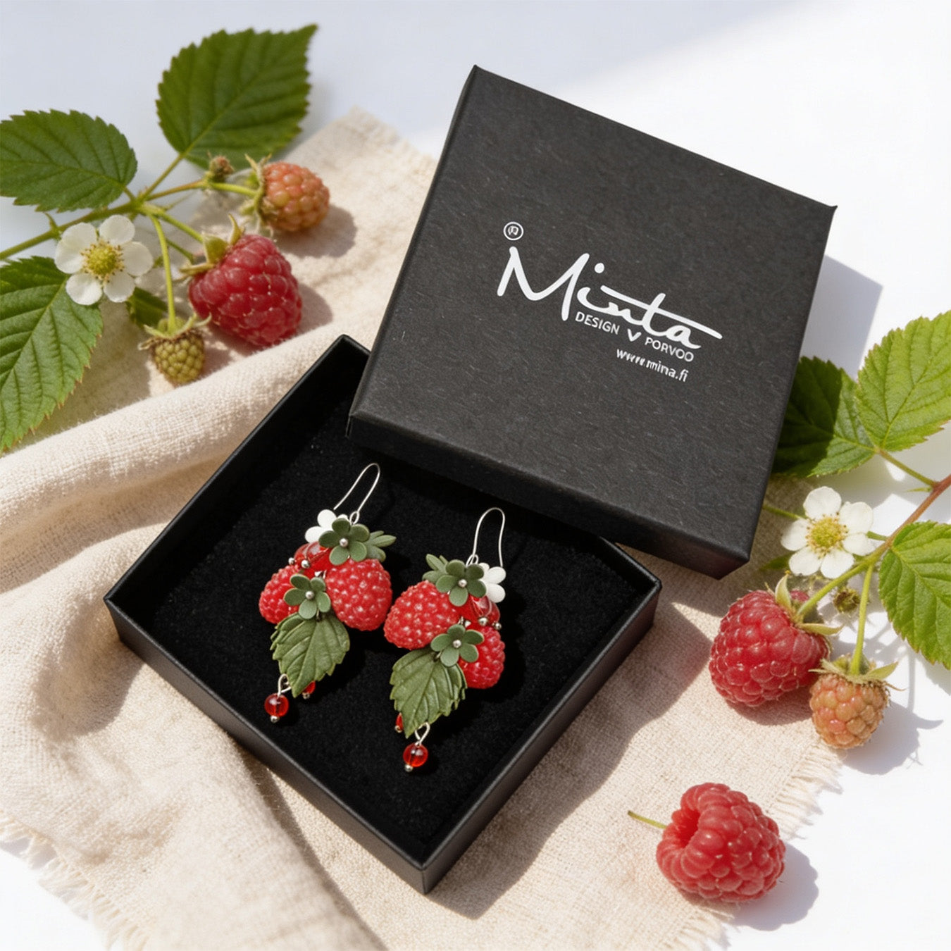 Wild Raspberry Earrings presented in a gift box, surrounded by natural linen fabric, fresh raspberries, raspberry leaves, and blossoms. Handmade in Finland by byMinta.