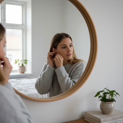 Woman putting on handmade Wild Raspberry stud earrings in front of a mirror. Small polymer clay berry earrings inspired by Nordic nature, handcrafted in Finland by byMinta.