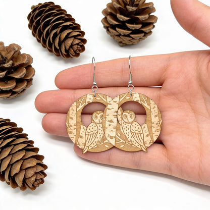 Handmade wooden owl earrings resting on a hand for size reference, surrounded by pine cones. Lightweight Finnish birch plywood earrings with detailed owl and birch forest engraving in soft natural light.