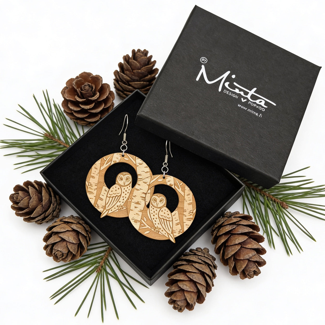 Handmade owl earrings displayed in a black jewelry gift box surrounded by pine cones and evergreen branches. Lightweight Finnish birch plywood earrings with detailed forest engraving, styled in a cozy Nordic woodland atmosphere.