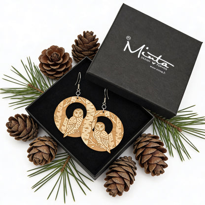 Handmade owl earrings displayed in a black jewelry gift box surrounded by pine cones and evergreen branches. Lightweight Finnish birch plywood earrings with detailed forest engraving, styled in a cozy Nordic woodland atmosphere.